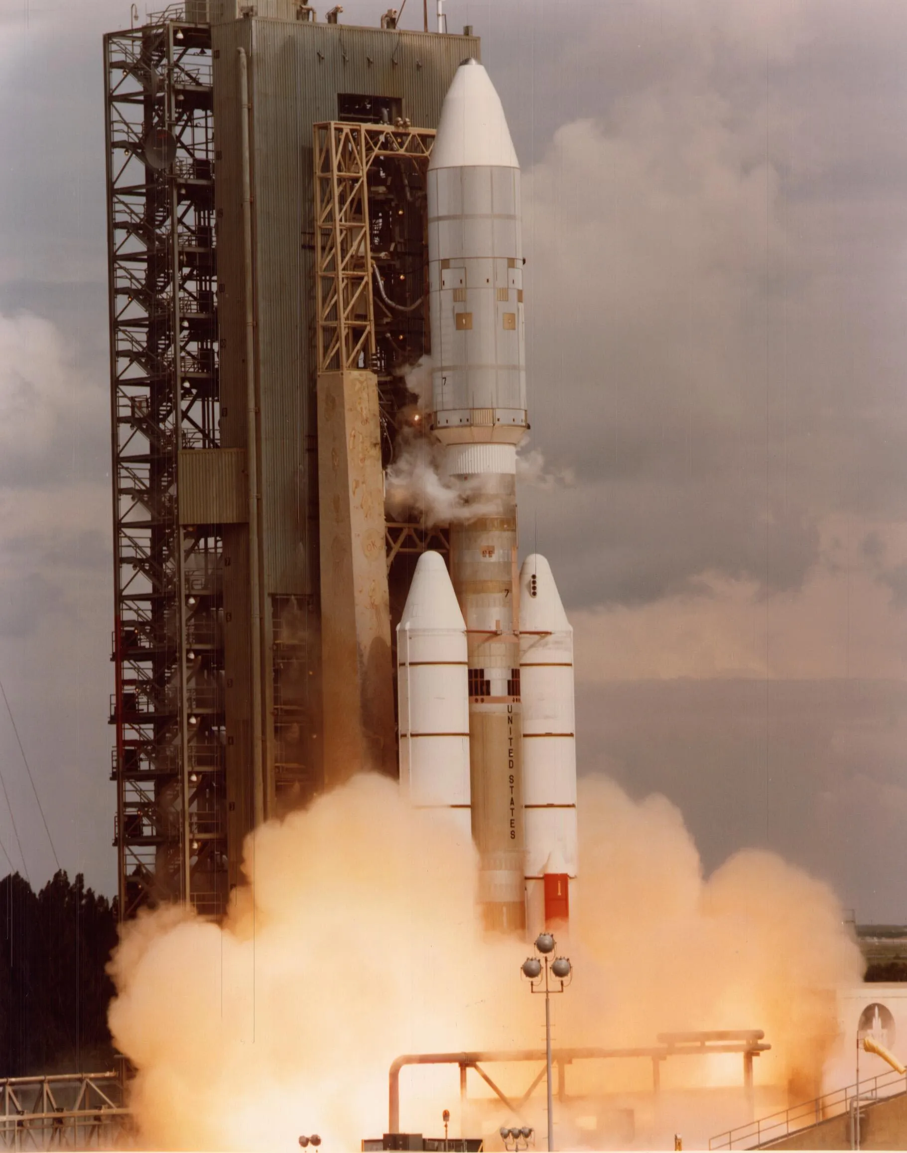 A Titan rocket with two white solid boosters lifts off beside a launch tower in a cloud of orange exhaust.