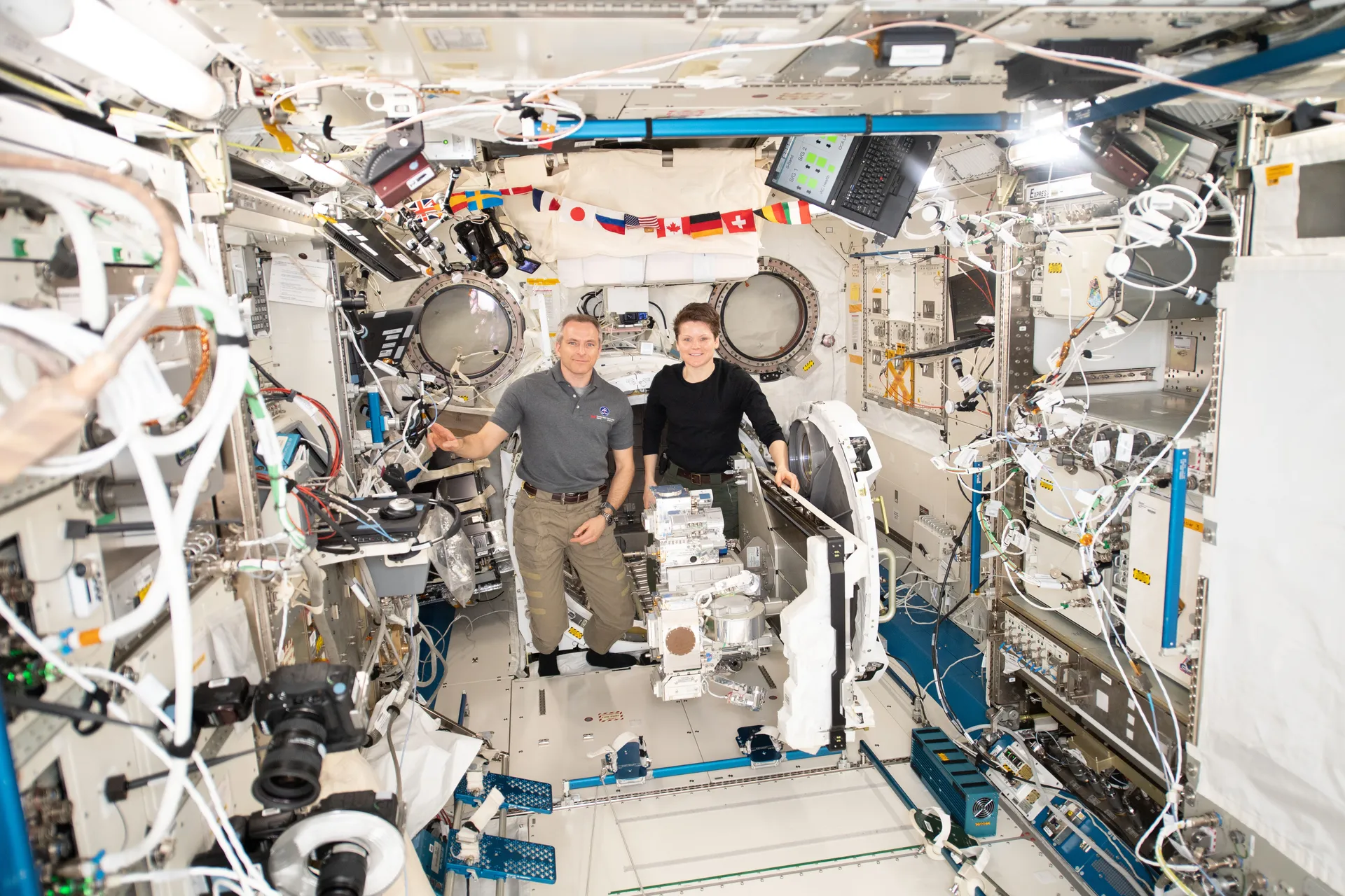 Two astronauts stand in a crowded space station module beside a white boxy experiment on an airlock slide table.