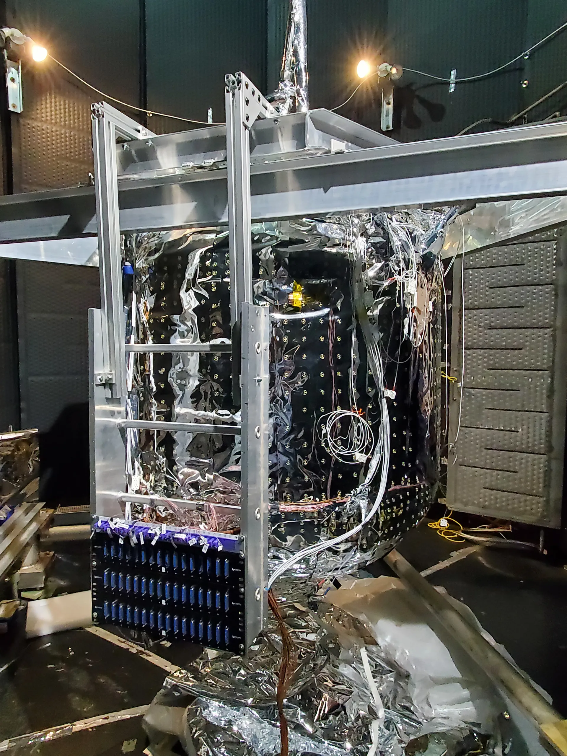 A tank covered in silver insulation and wired with sensors, mounted in a metal frame inside a dark vacuum chamber.