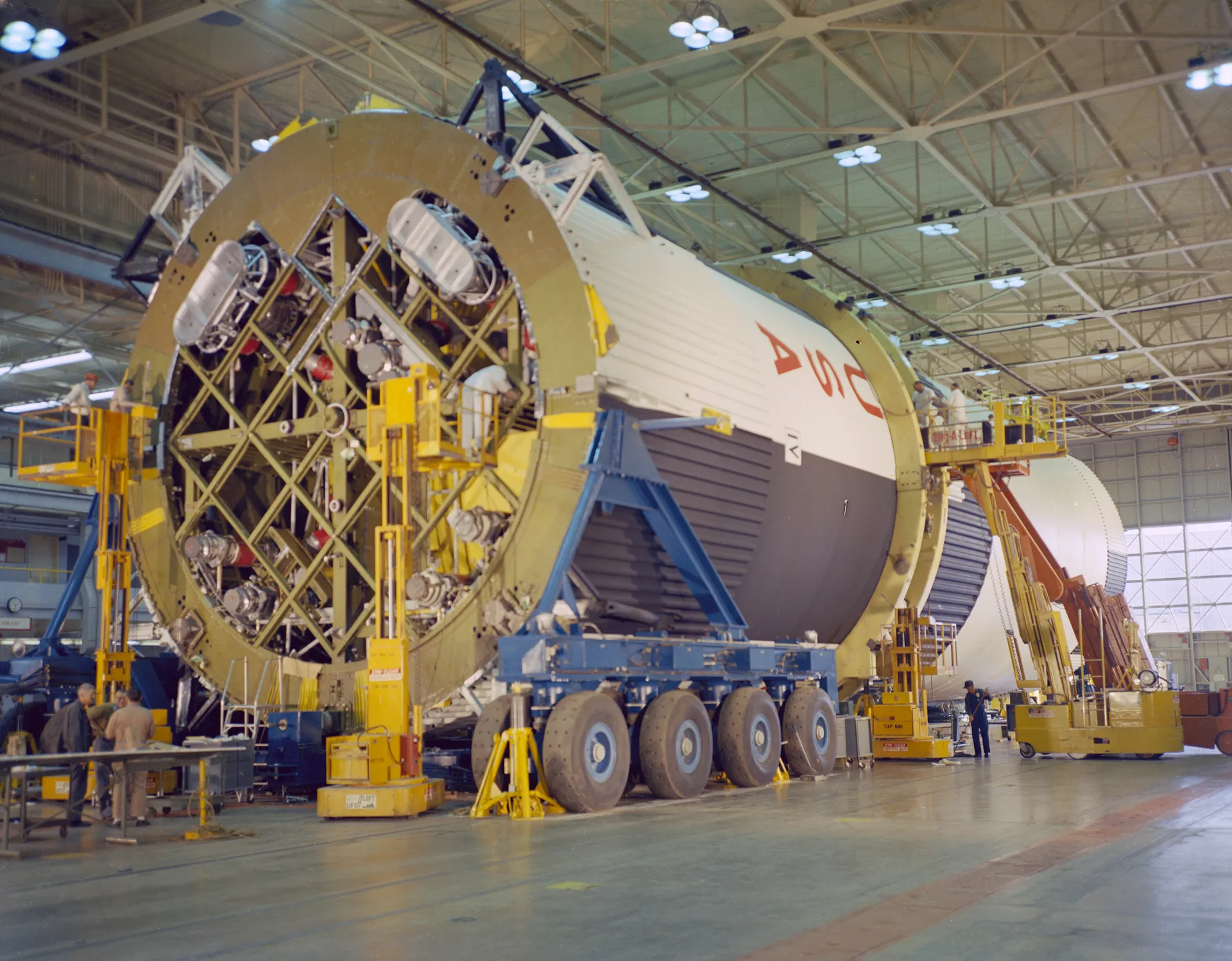 A huge white cylindrical rocket stage lies on its side on wheeled transporters in a factory hall, its base a lattice of thrust structure with workers and lifts around it.
