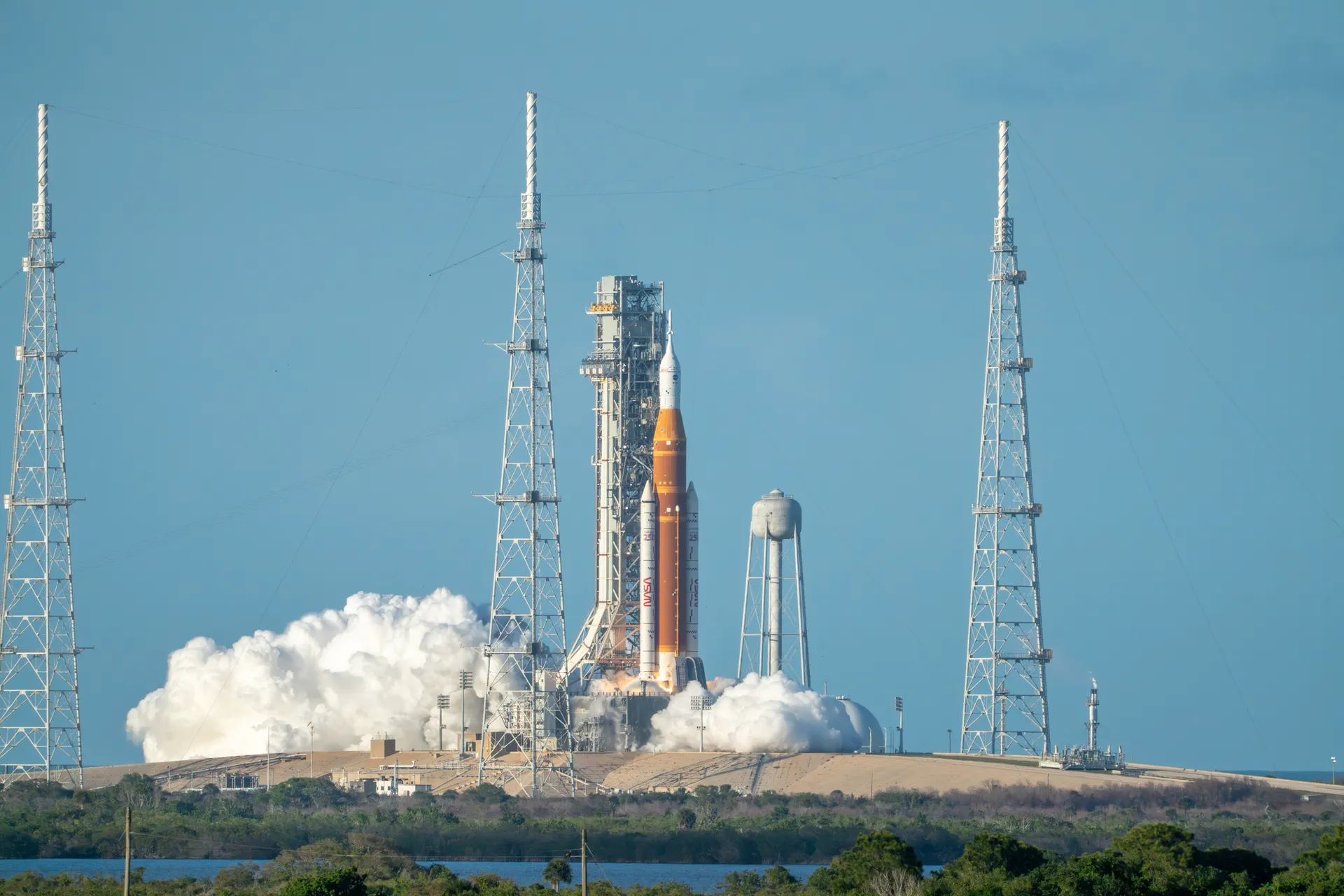 Seen across scrubland, SLS stands between three tall lightning towers at Launch Complex 39B as white exhaust spreads from the pad at ignition.