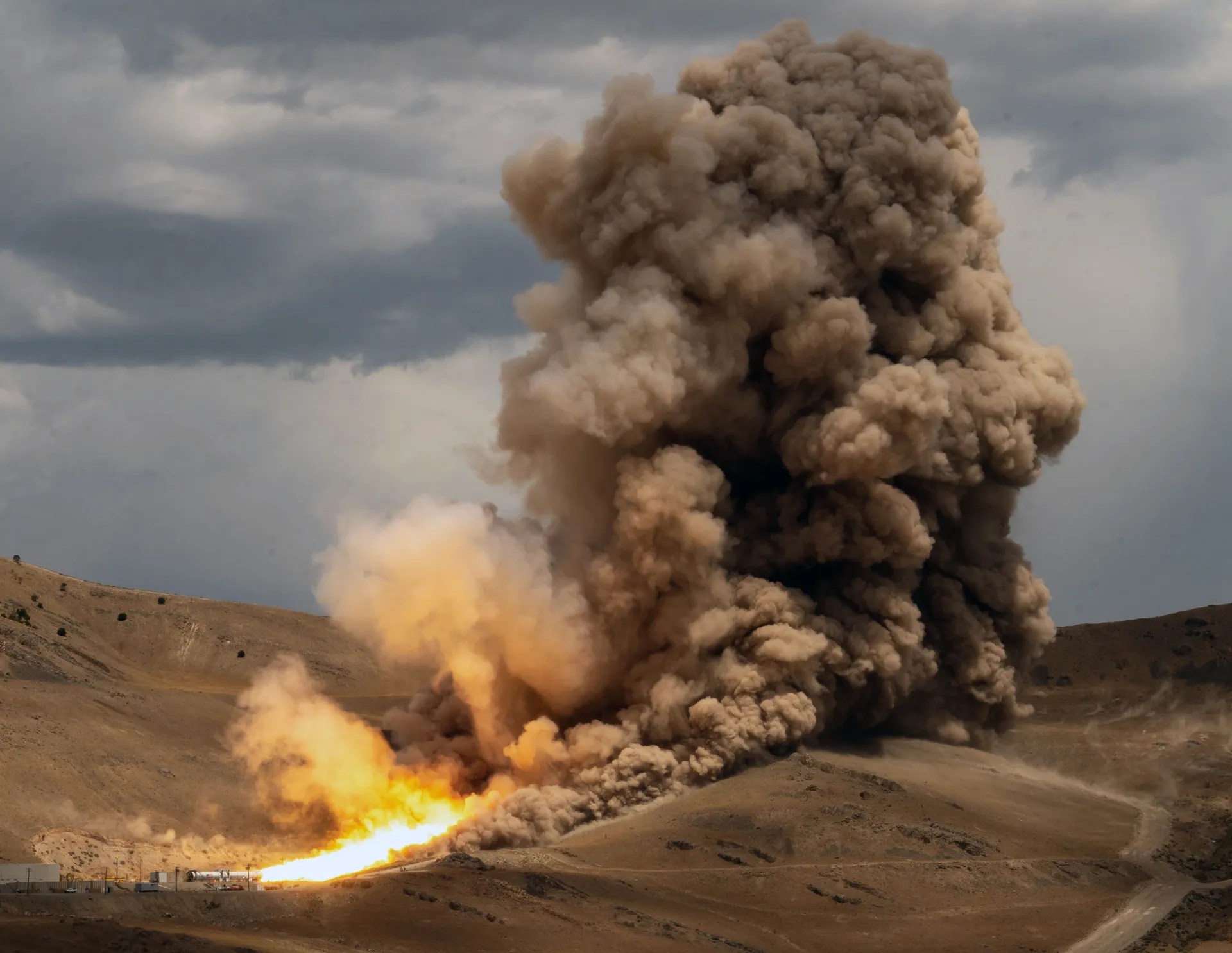 A horizontal solid rocket motor fires in a desert canyon, sending a long orange flame and a towering column of brown smoke into a cloudy sky.