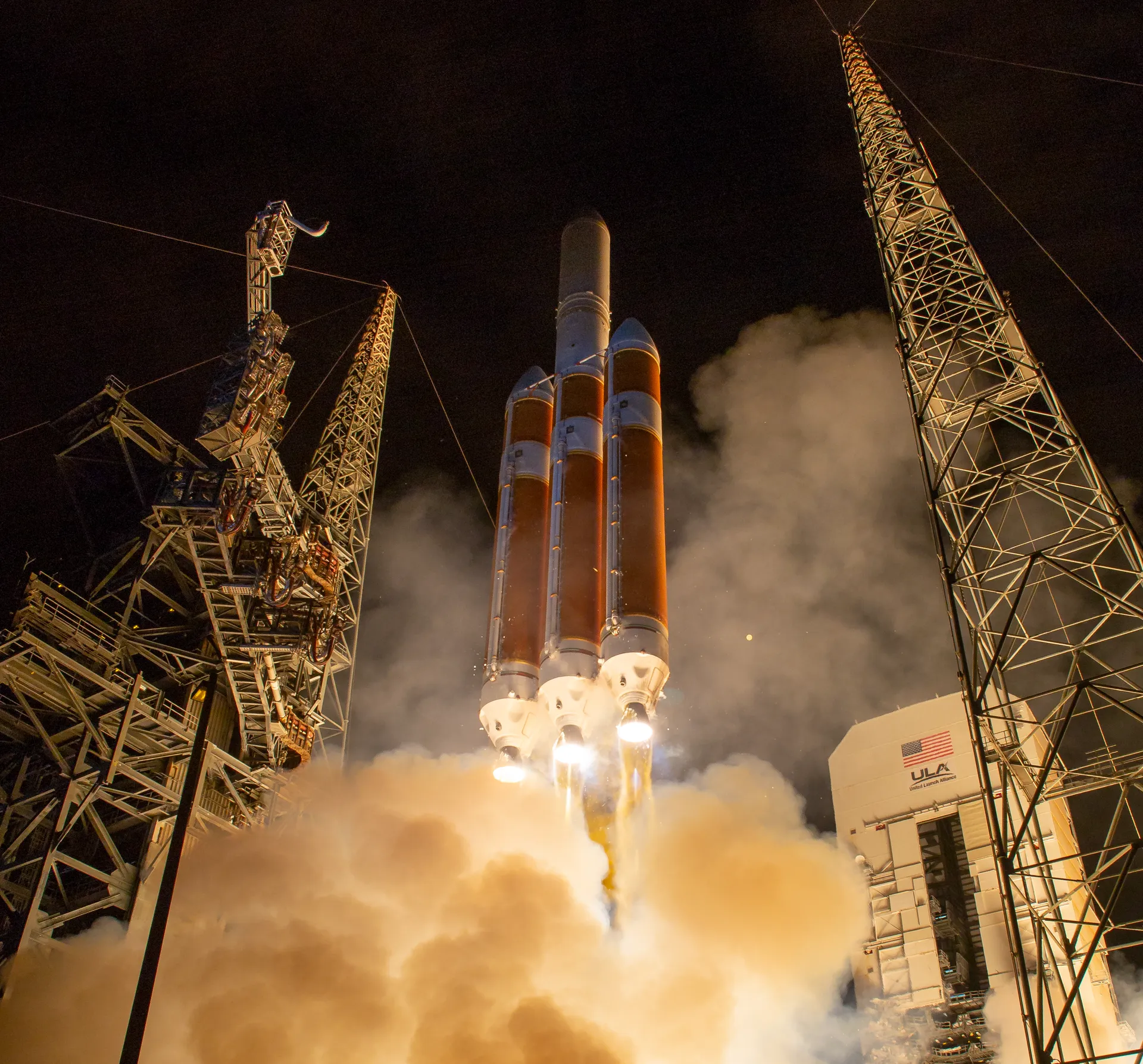A Delta IV Heavy rocket, three orange cores side by side, lifts off at night between two lattice towers in a glow of exhaust and smoke.