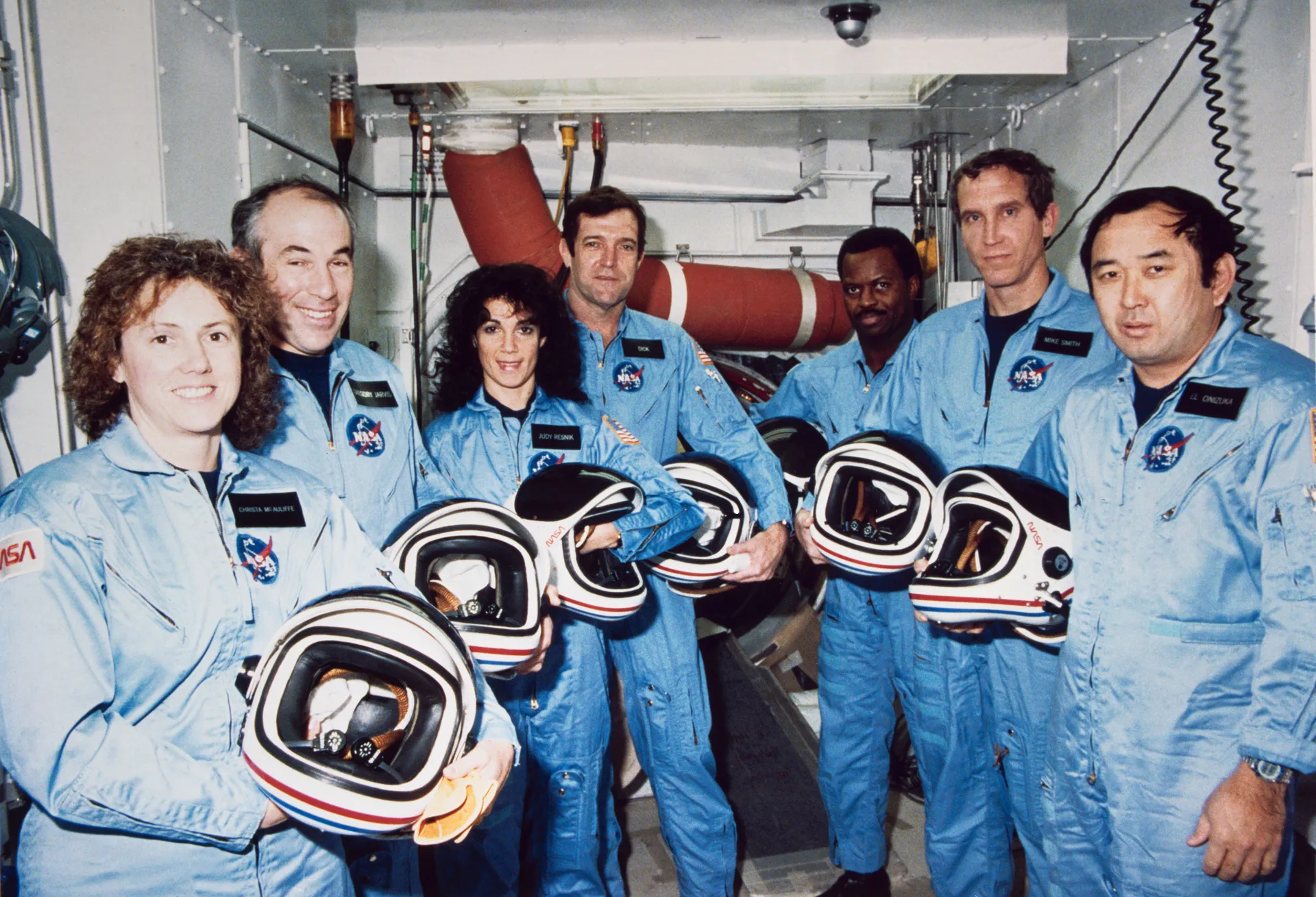 The seven STS-51L astronauts in blue flight suits, holding their helmets, in the White Room at the launch pad.