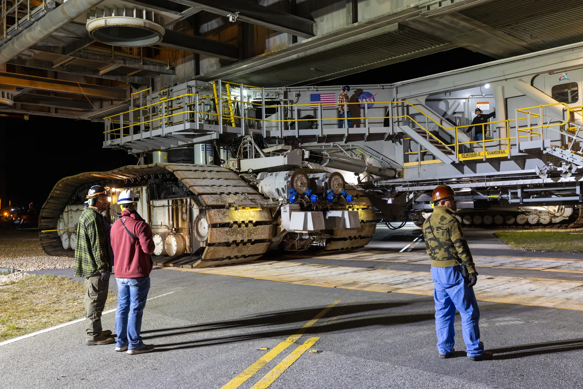 At night, workers in hard hats walk beside the enormous tracked treads of a crawler-transporter lit by floodlights on the crawlerway.