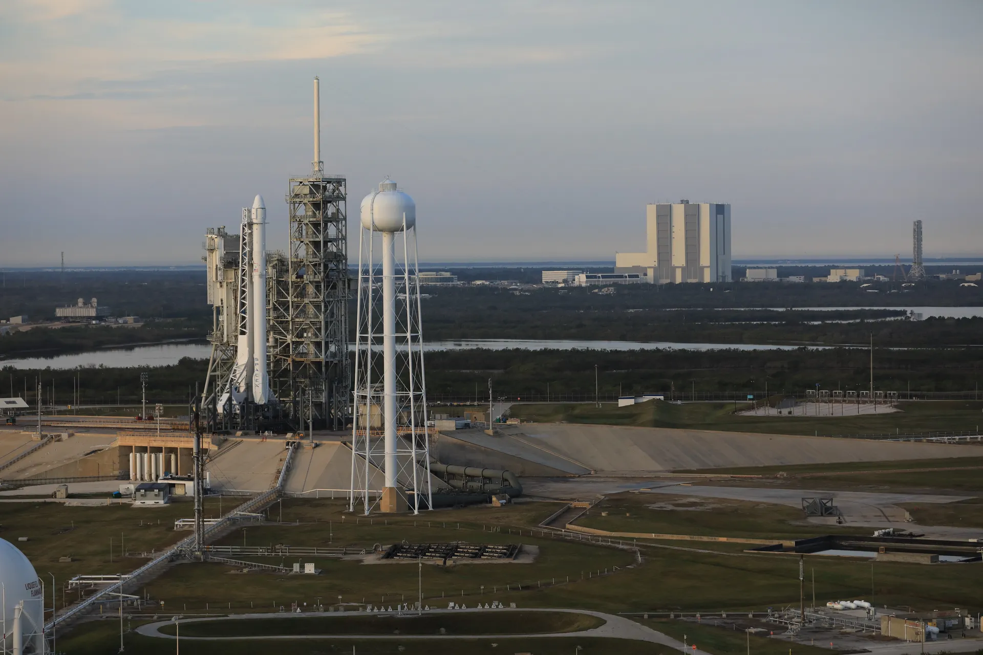 Aerial view of a Falcon 9 standing at Launch Complex 39A, with the Vehicle Assembly Building and the SLS mobile launcher in the distance across the marsh.