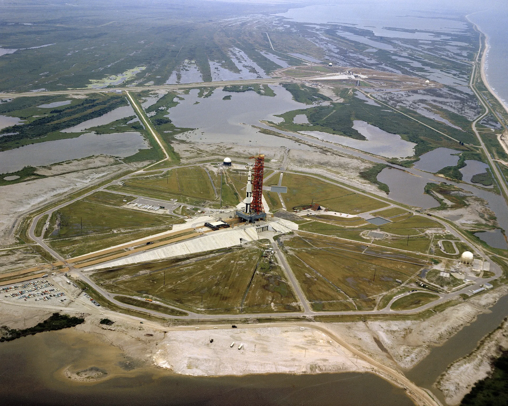 Aerial view of Launch Complex 39A in 1966: an octagonal pad with a Saturn V test vehicle and tower at the center, a straight crawlerway leading inland across marshland.