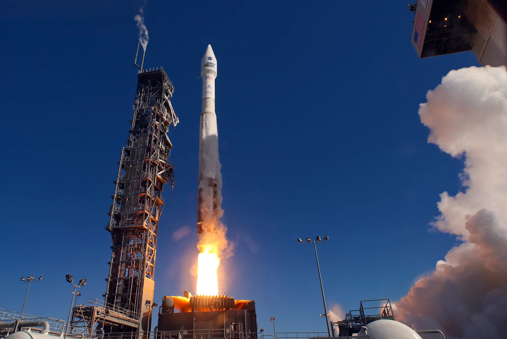 An Atlas V lifts off beside its launch tower under a deep blue sky, white exhaust billowing to the right.