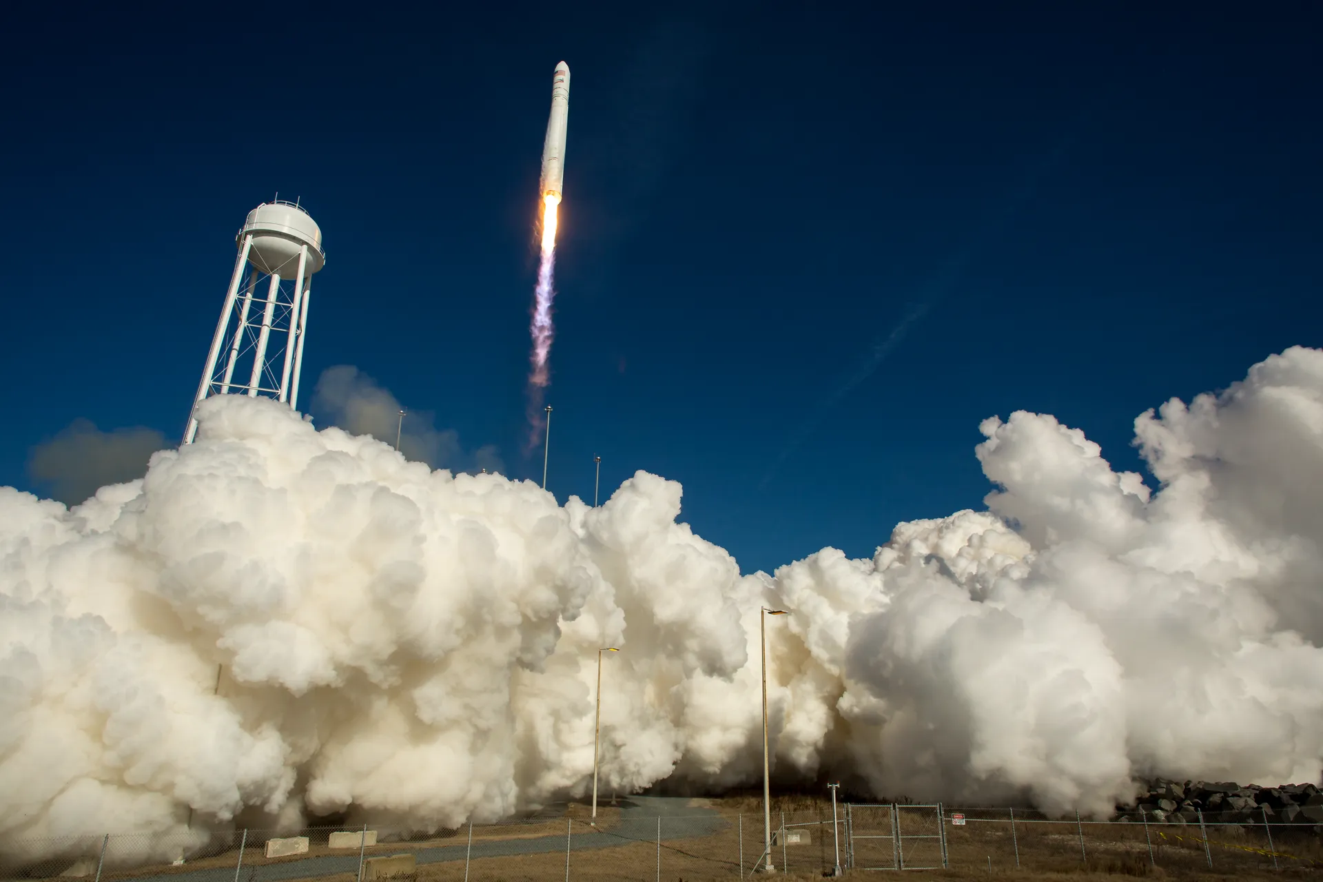 An Antares rocket climbs from Pad-0A at Wallops on a bright flame, a cloud of white exhaust spreading across the ground beside a water tower.