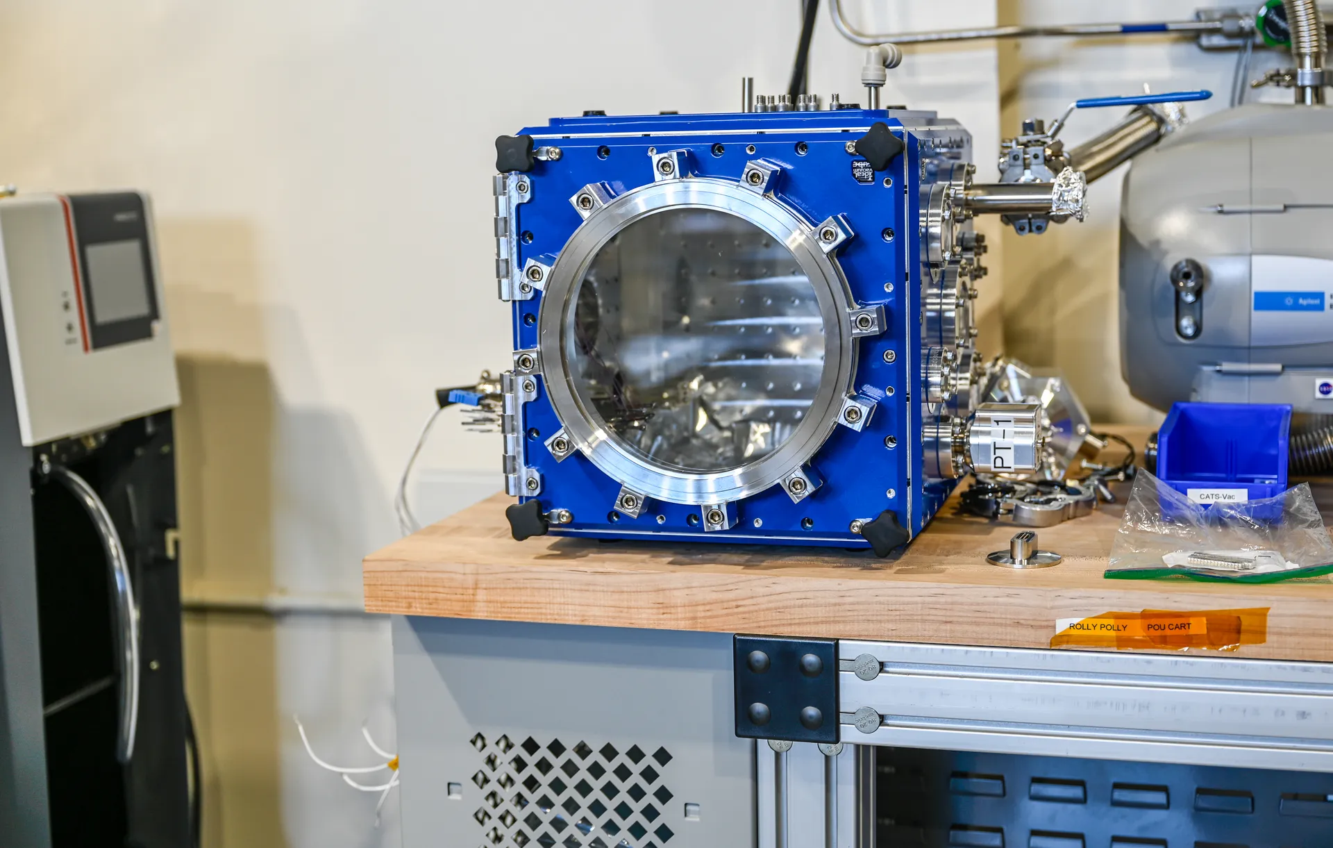 A blue cube-shaped vacuum chamber with a round window sits on a workbench among instruments.