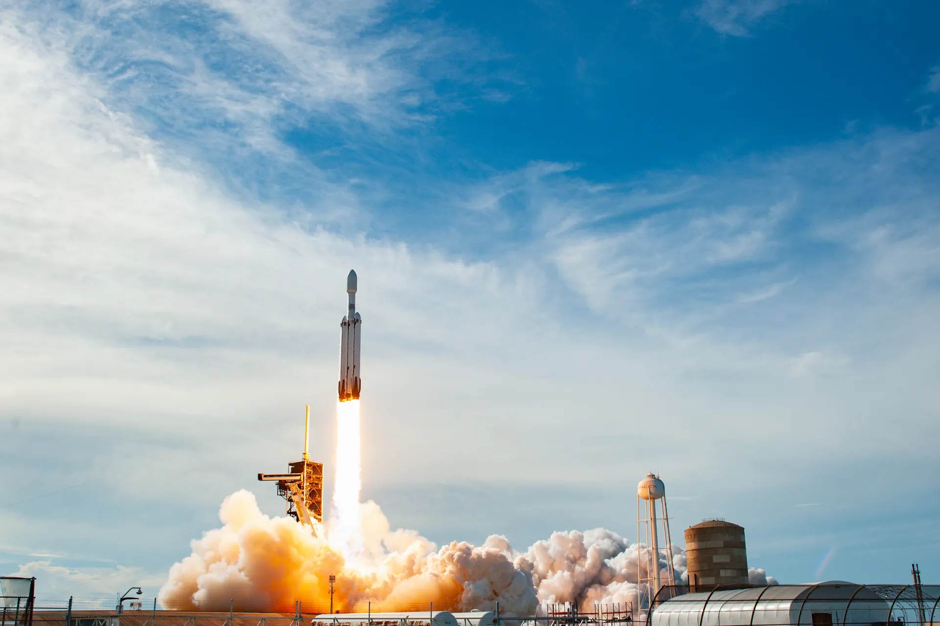 A Falcon Heavy rocket, three cores side by side, rises from the pad on a column of flame and steam under a blue sky with high clouds.