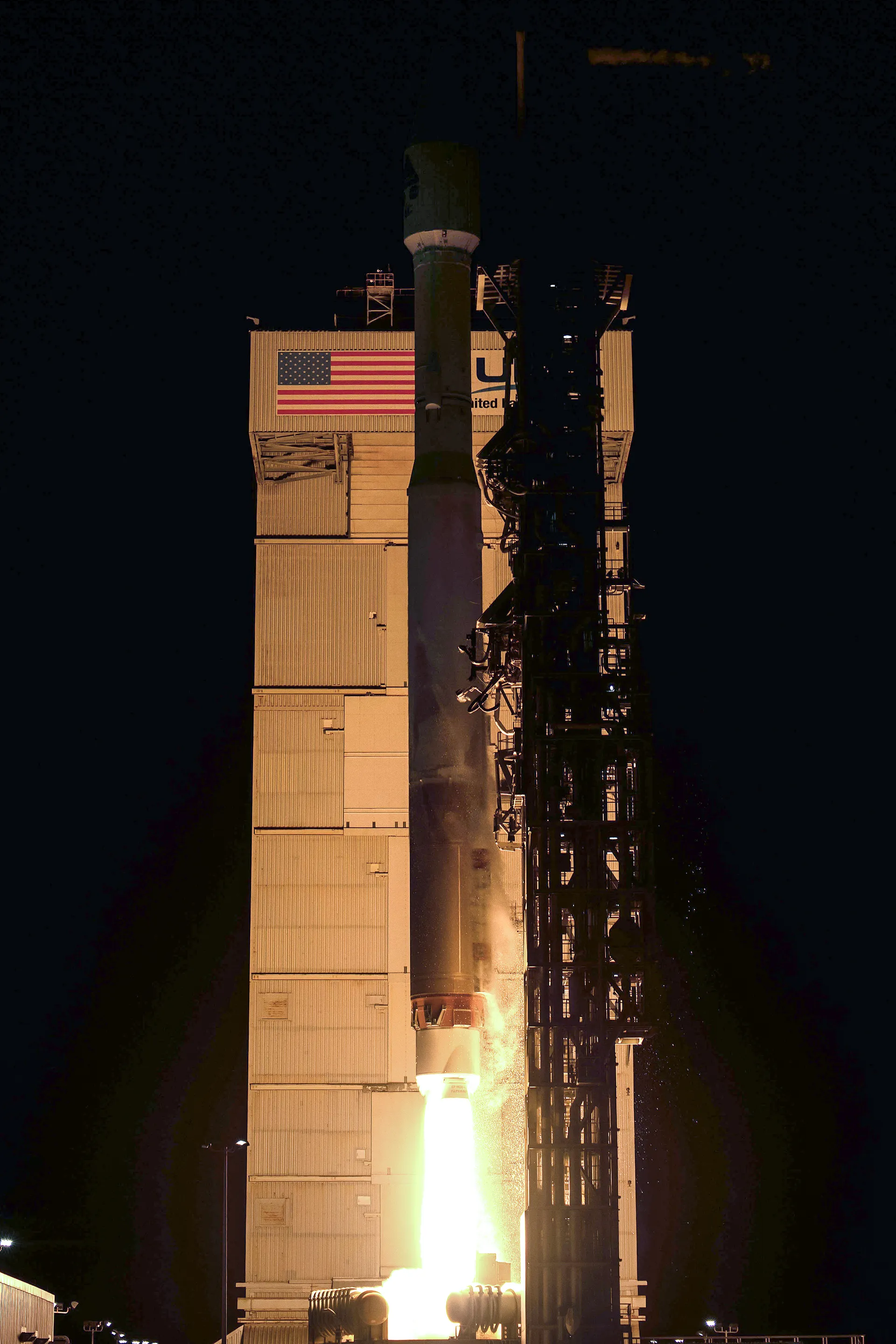 An Atlas V rocket rises from its pad at night beside the floodlit launch tower, its engine plume lighting the steel structure.
