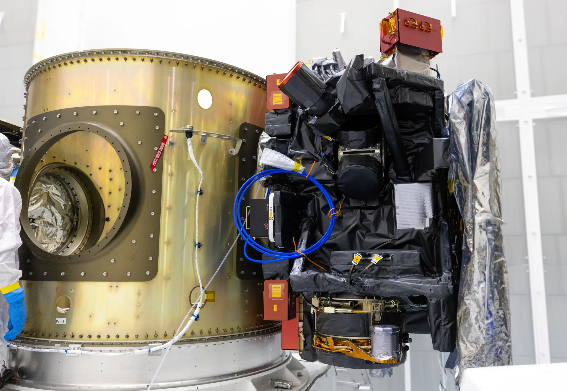 A boxy spacecraft wrapped in black insulation, with red protective covers on its instruments, mounted on the side of a large gold-colored ring in a clean room.